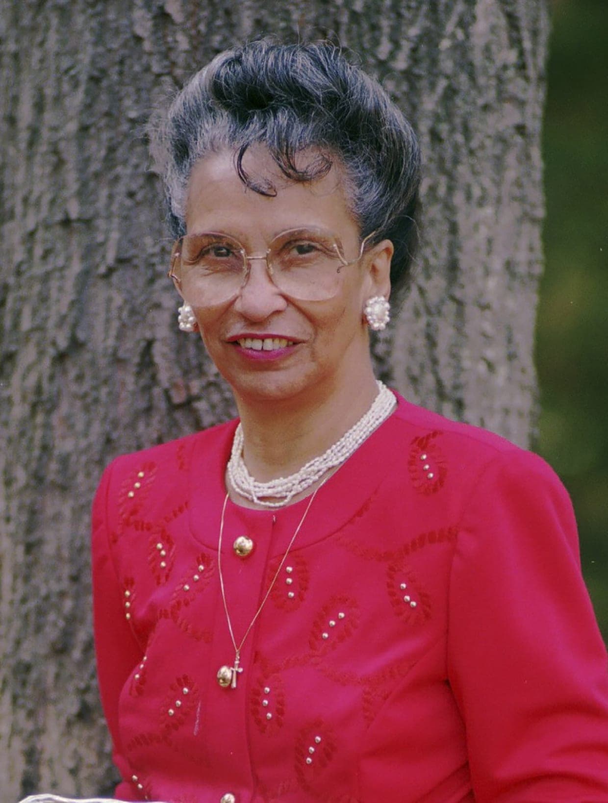 Jean W. Toomer, who founded MCRC in 1993, an older Black woman with softly greying upswept hair and wire-rimmed glasses, smiling, wearing a red jacket and pearls, photographed outdoors against a tree